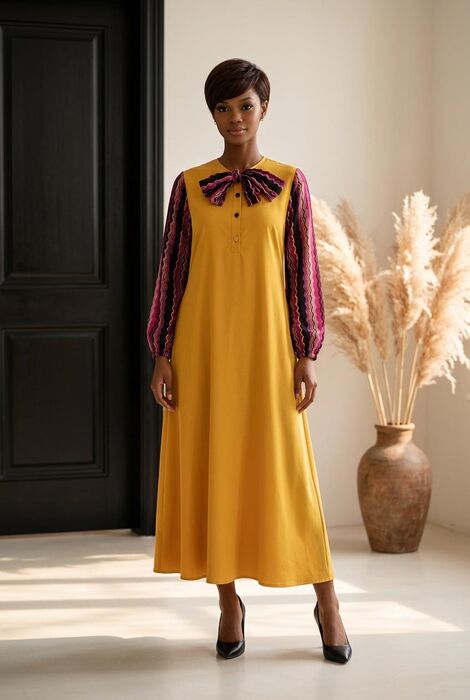A long-sleeved dress featuring a solid mustard yellow bodice and skirt with a round neckline. The sleeves are patterned with vertical wavy stripes in shades of pink, purple, and bl