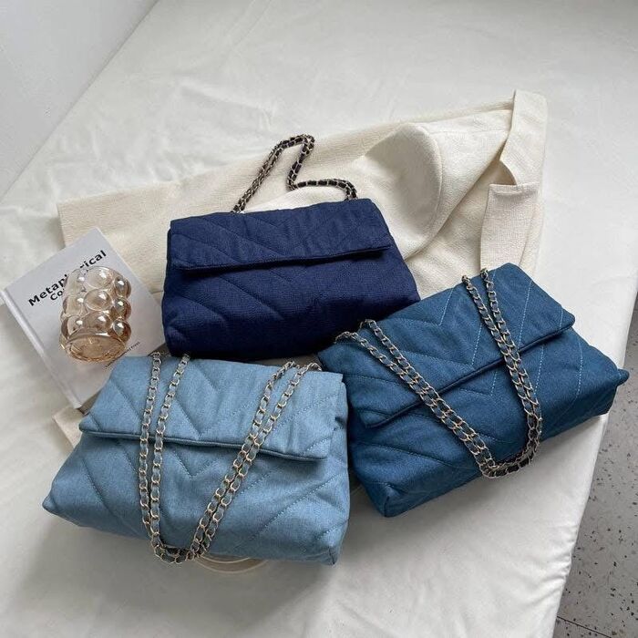Three quilted denim shoulder bags in varying shades of blue, ranging from light to dark. Each bag features a fold-over flap, a chevron-stitched pattern, and a silver-toned chain st
