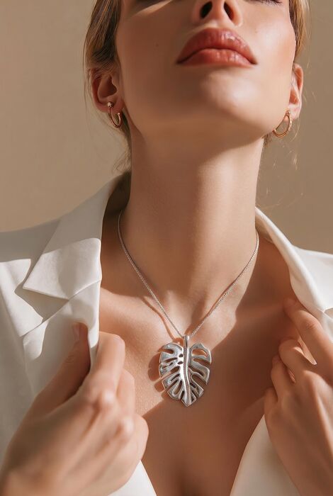 A silver-toned necklace featuring a large, detailed monstera leaf pendant suspended from a delicate chain. The pendant has a polished, reflective surface with cut-out sections that