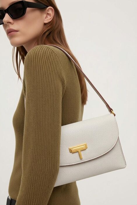 A white textured leather shoulder bag featuring a gold-toned T-shaped metal clasp on the front flap. The bag has a slim profile and a thin brown leather shoulder strap attached wit